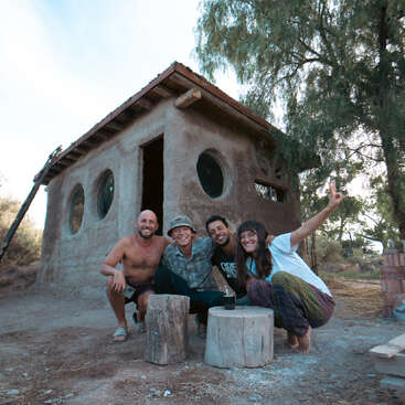Cuatro personas sonrientes posan felices delante de una casita rústica con ventanas redondas, rodeada de árboles, sentadas en taburetes de madera, disfrutando de un día soleado.