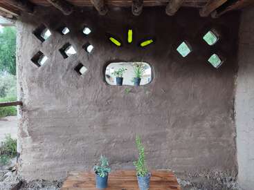 Cette image montre un mur rustique en terre crue avec des fenêtres carrées, des décorations réalisées à partir de bouteilles en verre recyclées, une fenêtre avec des plantes en pot et une table en bois avec deux autres plantes.