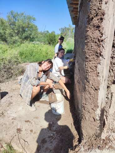 Trois personnes travaillent ensemble pour appliquer un enduit de boue sur un mur rustique à l'extérieur. Elles sourient, profitant de la journée ensoleillée, entourées de verdure et d'arbres.