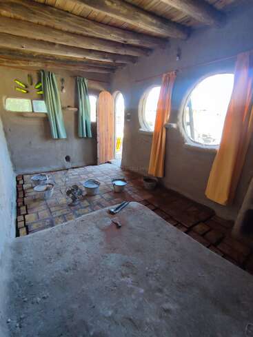 Cette pièce rustique est dotée d'un plafond en bois, de fenêtres rondes ornées de rideaux colorés, de murs en terre cuite, d'un sol en briques et de divers seaux et outils, créant ainsi une ambiance chaleureuse et naturelle.