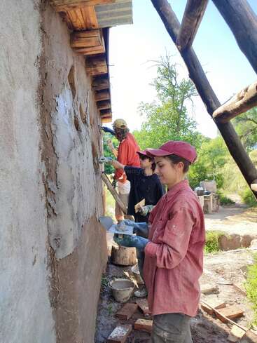 Three people work together plastering a cob wall with natural materials. They wear gloves and hats, applying the mixture carefully, surrounded by nature and construction materials.