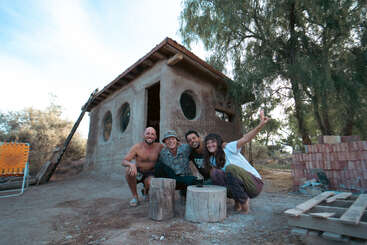 Four smiling people pose happily in front of a small rustic house with round windows, surrounded by trees, sitting on wooden stools, enjoying a sunny day.