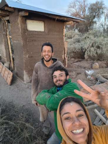 Three smiling friends pose for a selfie at a construction site, wearing dusty clothes. One flashes a peace sign, showing camaraderie and joy amid hard work outdoors.