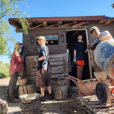 Four people work together on a construction project, possibly building with earth or cob. They smile, surrounded by buckets, a wheelbarrow, and an unfinished structure.