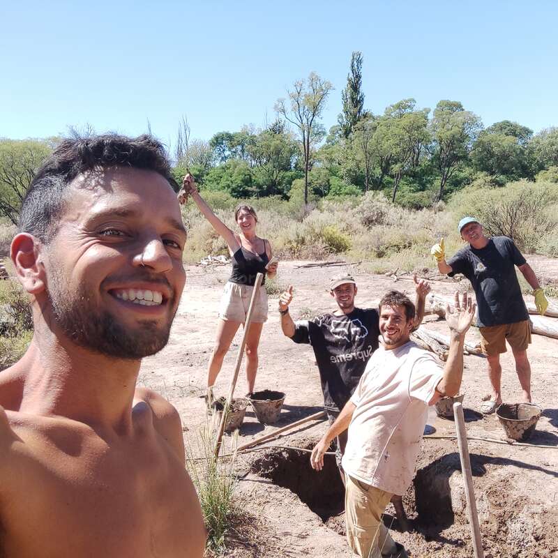 Eine Gruppe von fünf Personen lächelt und posiert, während sie in einer sonnigen, ländlichen Gegend mit Bäumen und Büschen im Hintergrund ein Loch graben.