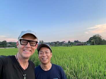 Two men are smiling and taking a selfie in front of a lush green field with a clear blue sky and distant trees in the background.