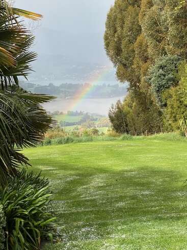 A vibrant green lawn is bordered by lush plants and tall trees, framing a distant view of a rainbow over a serene lake and landscape.