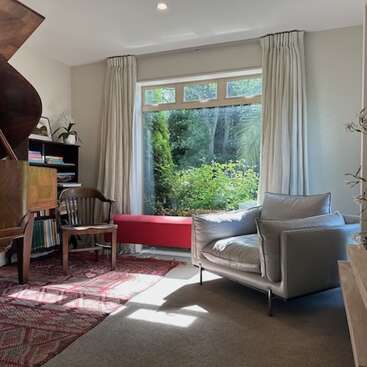 A cozy, sunlit room features a large window, piano, armchair, red rug, bookshelves, wooden chairs, and indoor plants, offering a serene view of the garden.