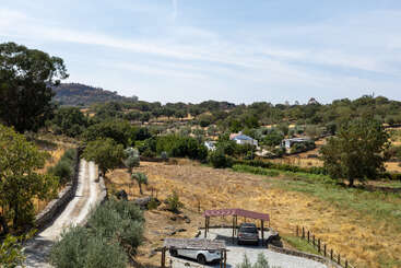 Uma paisagem rural apresenta uma longa e sinuosa estrada de terra, um carro estacionado sob uma garagem, campos de grama seca, árvores espalhadas e casas cercadas por colinas. Um campo tranquilo.