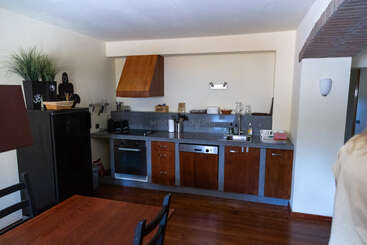 Esta imagem mostra uma cozinha moderna com armários de madeira, uma geladeira preta, forno, pia e bancada. Uma mesa de jantar com cadeiras está posicionada em primeiro plano.