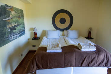 Esse quarto aconchegante tem duas camas de solteiro com lençóis brancos e cobertores marrons, arte na parede, luminárias de cabeceira, toalhas e uma peça decorativa redonda acima da cabeceira.