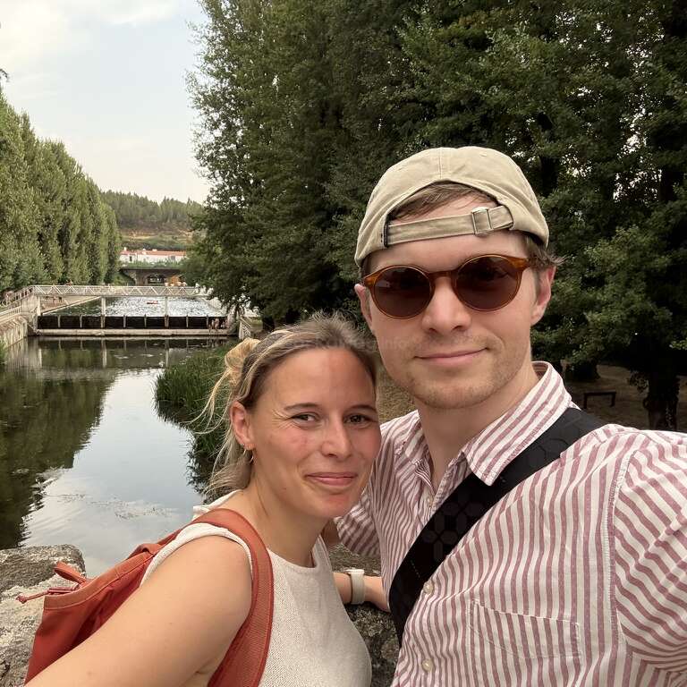 Um casal sorridente tira uma selfie ao ar livre perto de um rio calmo, cercado por altas árvores verdes, usando roupas casuais de verão. O plano de fundo inclui uma pequena ponte.