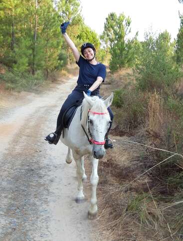 Une personne chevauchant joyeusement un cheval blanc sur un sentier forestier, souriante, portant un casque et des gants, levant un bras en l'air, entourée d'arbres verts.