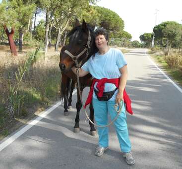 La imagen muestra a una mujer de pie en un camino con un caballo marrón, vestida con camisa y pantalones azul claro, con árboles y hierba de fondo.