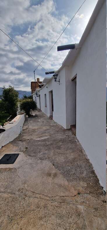 A rustic walkway runs beside a whitewashed building with outdoor lamps. Cracked concrete path, greenery along the side, cloudy sky, and mountains visible in the background.