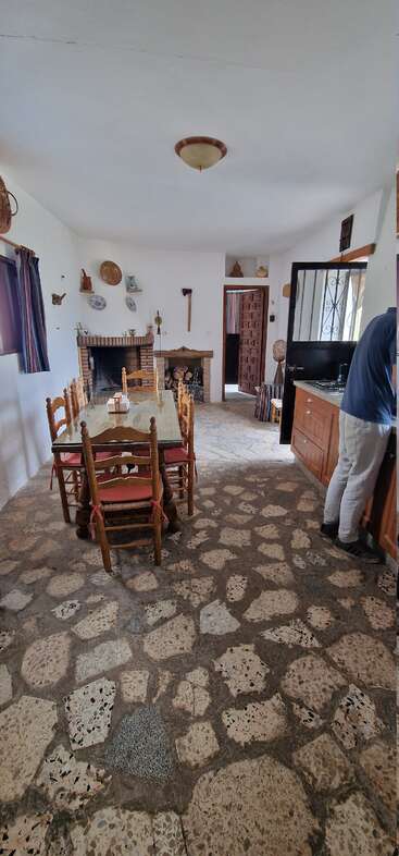 A rustic dining room with a glass-topped table, fireplace, stone floor, shelves with pottery, and a man standing near the wooden kitchen counter on the right.