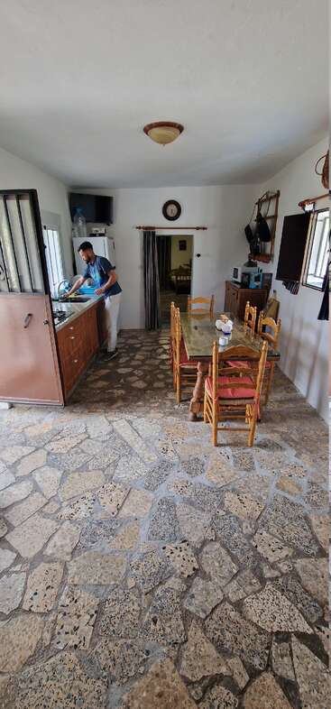 A man washes dishes in a rustic kitchen with stone floor, wooden chairs, a glass table, and basic appliances. A cozy, simple, country-style interior.