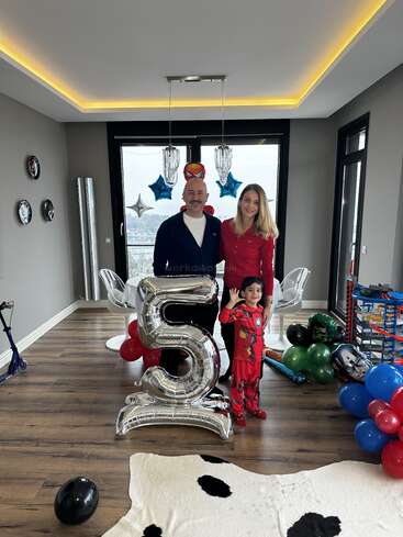 A family celebrates a child's fifth birthday at home. The child wears an Iron Man costume. Large balloons, superhero decorations, and toys fill the festive room.