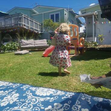 Uma menina com um vestido floral caminha descalça na grama, carregando uma bolsa, em um quintal ensolarado com brinquedos, assentos e decorações festivas.