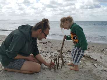 Um homem e uma criança estão construindo uma estrutura com gravetos em uma praia de areia, desfrutando de um momento lúdico e criativo perto da água calma do oceano.