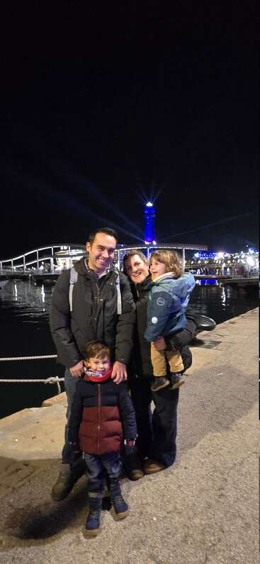 Eine lächelnde vierköpfige Familie posiert nachts am Ufer. Hinter ihnen funkeln eine Brücke und ein Turm mit blauen und weißen Lichtern und sorgen für eine festliche Stimmung.
