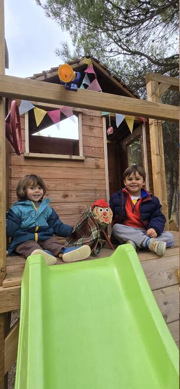 Zwei fröhliche Kinder sitzen auf einer Holzplattform, die mit bunten Wimpeln geschmückt ist. Eine grüne Rutsche führt nach unten. Sie halten eine Figur fest, die in eine karierte Decke gewickelt ist.