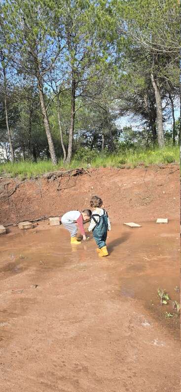 Zwei Kinder in gelben Stiefeln spielen in einer matschigen Pfütze, umgeben von roter Erde und grünem Wald, und genießen an einem sonnigen Tag draußen die Natur und das Entdecken.