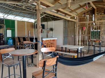 Esta rústica cafetería al aire libre cuenta con mesas y sillas de madera, un bar, dispensador de agua, ventiladores, una hamaca y paredes de metal corrugado, que crean un ambiente relajado e informal.