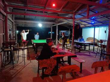 Un bar al aire libre con muebles de madera. La gente juega al billar y socializa, mientras otros se sientan en mesas bajo luces de colores, creando un ambiente relajado e informal.