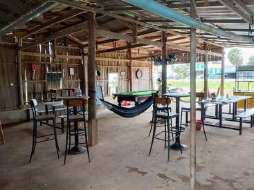 Esta cabaña rústica de madera al aire libre cuenta con mesas, taburetes, una hamaca, una mesa de billar y una decoración informal, que crean un ambiente relajado y social con vistas al exterior.
