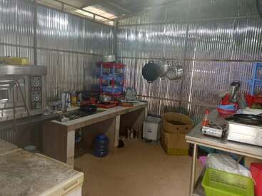 Esta imagen muestra una cocina sencilla con paredes de metal corrugado, electrodomésticos básicos, utensilios, ollas y diversos suministros organizados en estanterías, mostradores y estantes colgantes.