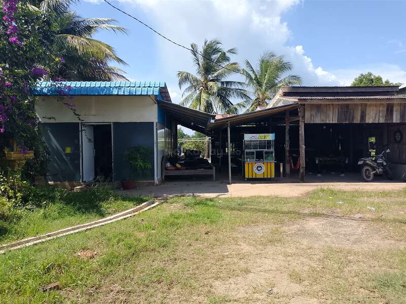 A rural scene featuring two small rustic buildings, palm trees, a food stall cart, a motorcycle, open grassy area, and bright sunlight under a clear blue sky.