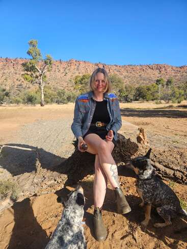 A woman sits on a tree stump outdoors, holding a ball, with two attentive dogs nearby. Rocky hills, trees, and blue sky are in the background.