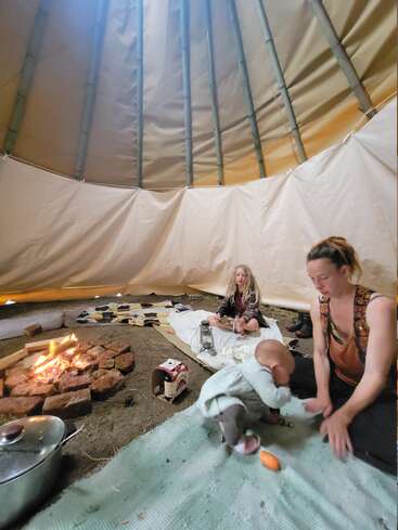 Inside a tipi, a woman, child, and baby gather around a central fire. Cozy blankets and personal items add warmth to this intimate, communal setting.