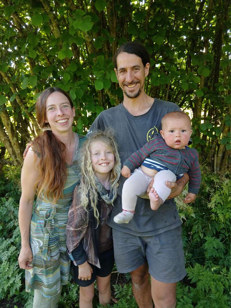 A happy family of four stands outdoors in front of leafy green trees, smiling warmly. The father holds a baby, while the mother and older child stand nearby.