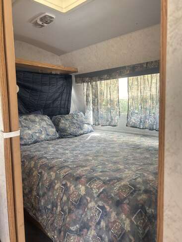 Essa imagem mostra um quarto pequeno e aconchegante dentro de um trailer ou camper. A colcha, os travesseiros e as cortinas têm padrões geométricos discretos que combinam entre si. A luz natural entra suavemente.