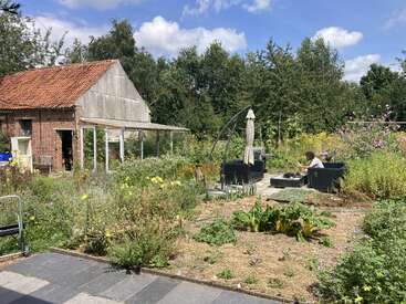 Eine rustikale Gartenszene mit einem alten Backsteinschuppen, wild wuchernden Pflanzen, Sitzgelegenheiten im Freien und einer Person, die sich an einem sonnigen Tag unter einem Sonnenschirm entspannt.