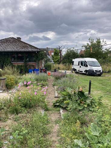 Ein lebhafter Bauerngarten mit blühenden Blumen, üppigem Grün und einem Steinweg. Ein weißer Lieferwagen steht in der Nähe unter einem dramatischen, bewölkten Himmel.