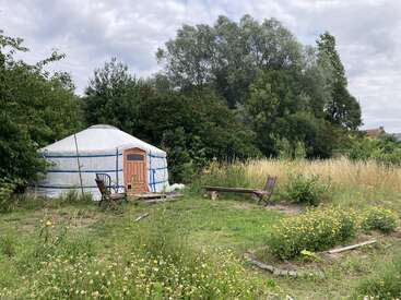 Eine weiße Jurte steht auf einer üppig grünen Wiese, umgeben von Bäumen, hohem Gras, Holzstühlen, einer Bank, Wildblumen und unter einem bewölkten Himmel.