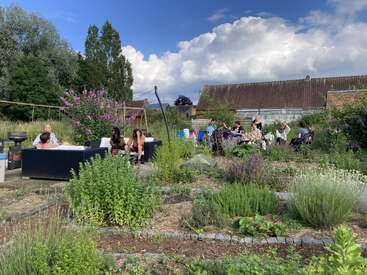 Eine Gruppe von Leuten chillt und unterhält sich in einem üppig grünen Gemeinschaftsgarten unter strahlend blauem Himmel, umgeben von Pflanzen, Blumen und rustikalen Gebäuden.