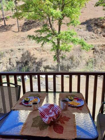 Une table en verre est dressée pour deux personnes avec des assiettes de petit-déjeuner, des tasses à café et une corbeille à pain sur un porche donnant sur un paysage sec, vallonné et arboré.