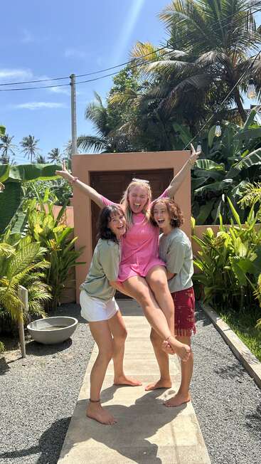 Tres mujeres jóvenes posan alegremente al aire libre en un día soleado, dos levantando a la tercera. Exuberantes plantas tropicales, palmeras y un cielo azul brillante las rodean. Momentos de alegría.