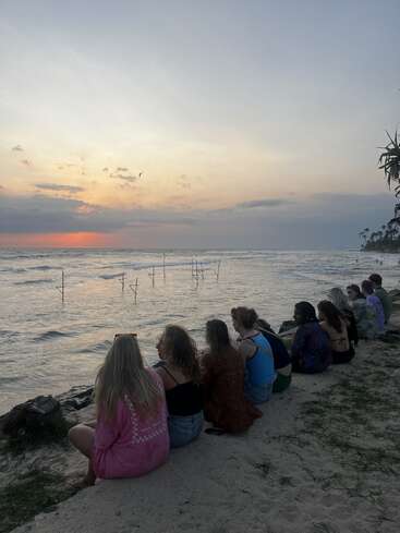 Eine Gruppe von Menschen sitzt an einem Sandstrand und beobachtet den ruhigen Ozean bei Sonnenuntergang. Sanfte Wellen, Angelruten und ein Vogel runden das friedliche Bild ab.