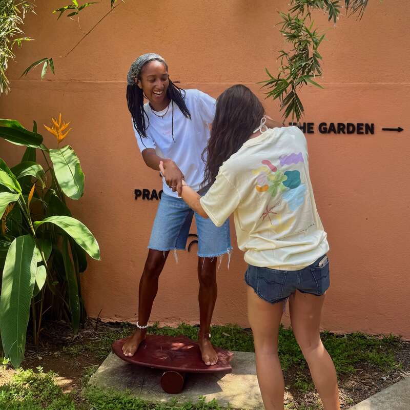 Dos amigos se divierten al aire libre, uno en equilibrio sobre una tabla mientras el otro le ayuda. Sonríen, rodeados de exuberante vegetación y una pared naranja. Un momento divertido.