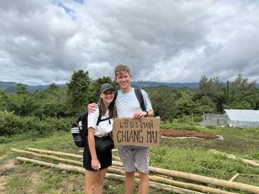 Una pareja sonriente se encuentra al aire libre, en medio de una exuberante vegetación, sosteniendo un cartel de cartón en el que se lee "Chiang Mai". Llevan ropa informal y mochilas, rodeados de montañas y cielos nublados.