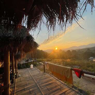 La imagen representa una serena puesta de sol desde una terraza de madera, con techo de paja y una barandilla adornada con toallas, sobre un pintoresco fondo montañoso.