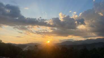 L'image montre un magnifique coucher de soleil sur des collines et des montagnes ondulantes. Les rayons du soleil percent les nuages et projettent une lumière dorée sur le paysage, créant une scène paisible et pittoresque.