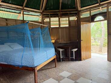 This cozy rustic room features a bed with a blue mosquito net, wooden walls, tiled floor, small table with stools, and an open door to nature.