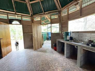 This rustic kitchen features wooden walls, green roof panels, tiled floors, open doors, a simple concrete sink, natural light, and a dog standing at the entrance.