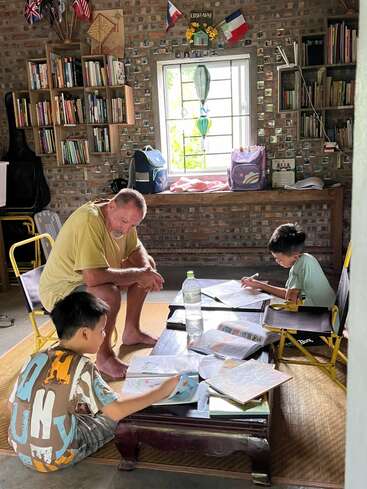 A man and two boys study together in a cozy room with bookshelves, flags, and a window. Notebooks, water bottle, and backpacks on the table.